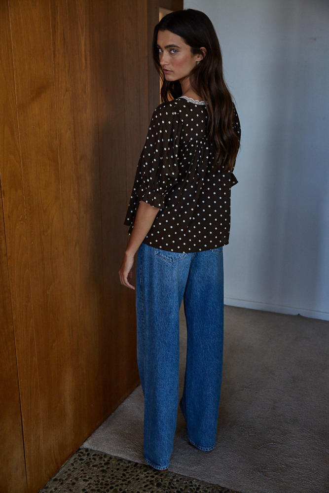 Woman wearing a black polka dot blouse and blue jeans standing against a wooden wall.