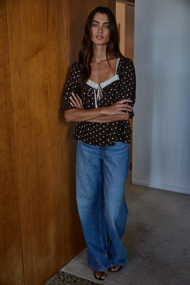 Woman wearing a polka dot top and blue jeans standing against a wooden wall.