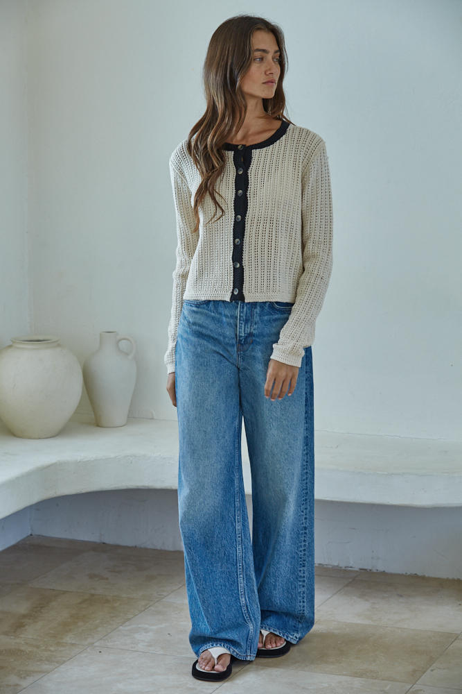 Woman wearing a cream cardigan and blue jeans standing in a minimalistic room.