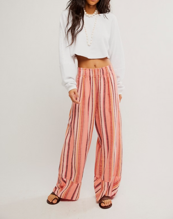 A model wearing wide-leg striped pants in shades of pink, orange, and beige, paired with a white cropped long-sleeve top and brown sandals, against a plain white studio background.