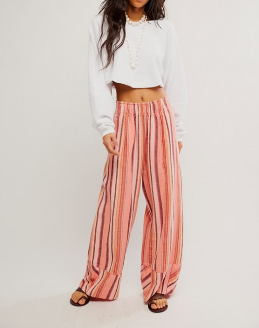 A model wearing wide-leg striped pants in shades of pink, orange, and beige, paired with a white cropped long-sleeve top and brown sandals, against a plain white studio background.