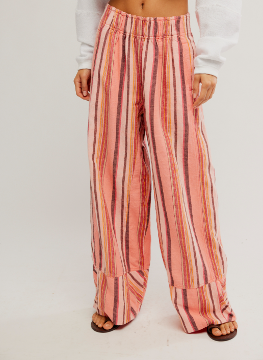 A person wearing loose, wide-leg striped pants in pink, peach, orange, and dark brown tones, paired with a white long-sleeve top and dark sandals, photographed against a plain light background.