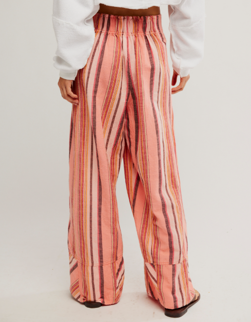 A person shown from the back wearing wide-leg striped pants in pink, orange, and brown tones, paired with a white long-sleeve top and dark shoes, against a plain light background.