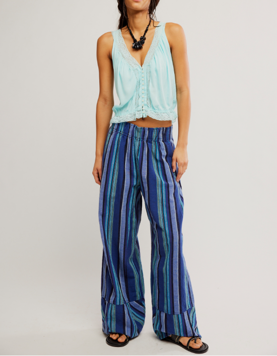 A model wearing a light blue sleeveless crop top and blue striped wide-leg pants, paired with black sandals and a black beaded necklace, against a plain light gray studio background.
