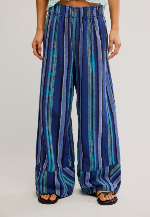 A person wearing wide-leg striped pants in shades of blue, teal, and navy, paired with black flip-flop sandals against a plain light gray studio background.