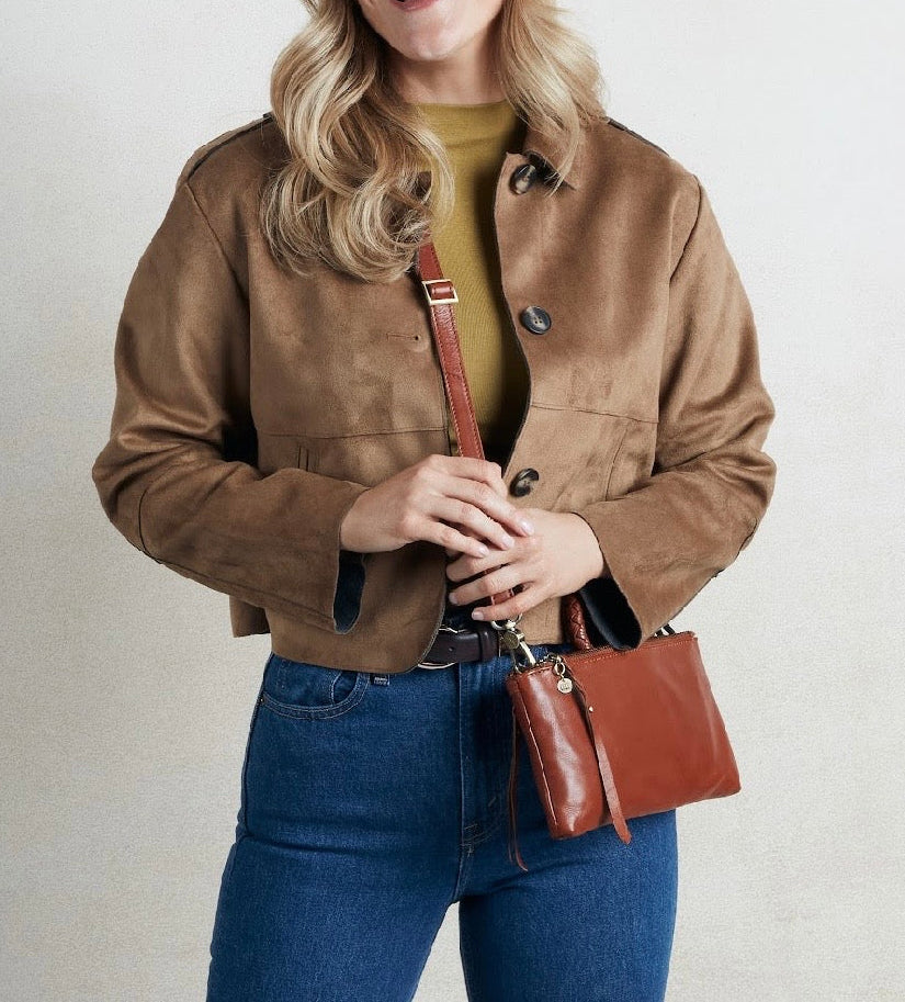 A woman wearing a brown suede jacket over a mustard yellow top and blue jeans, carrying a brown leather crossbody bag against a light gray background.