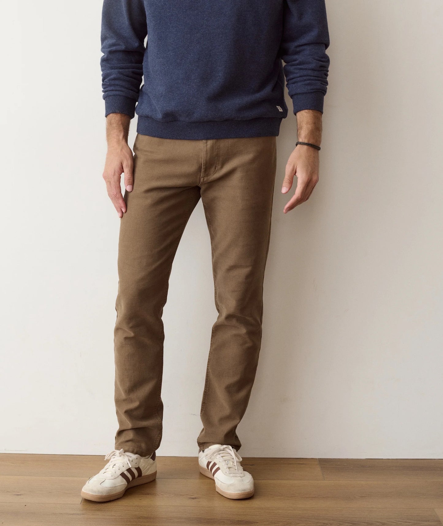 A person wearing slim brown pants, a navy blue long-sleeve top, and white low-top sneakers, standing against a plain off-white wall on a light wood floor.