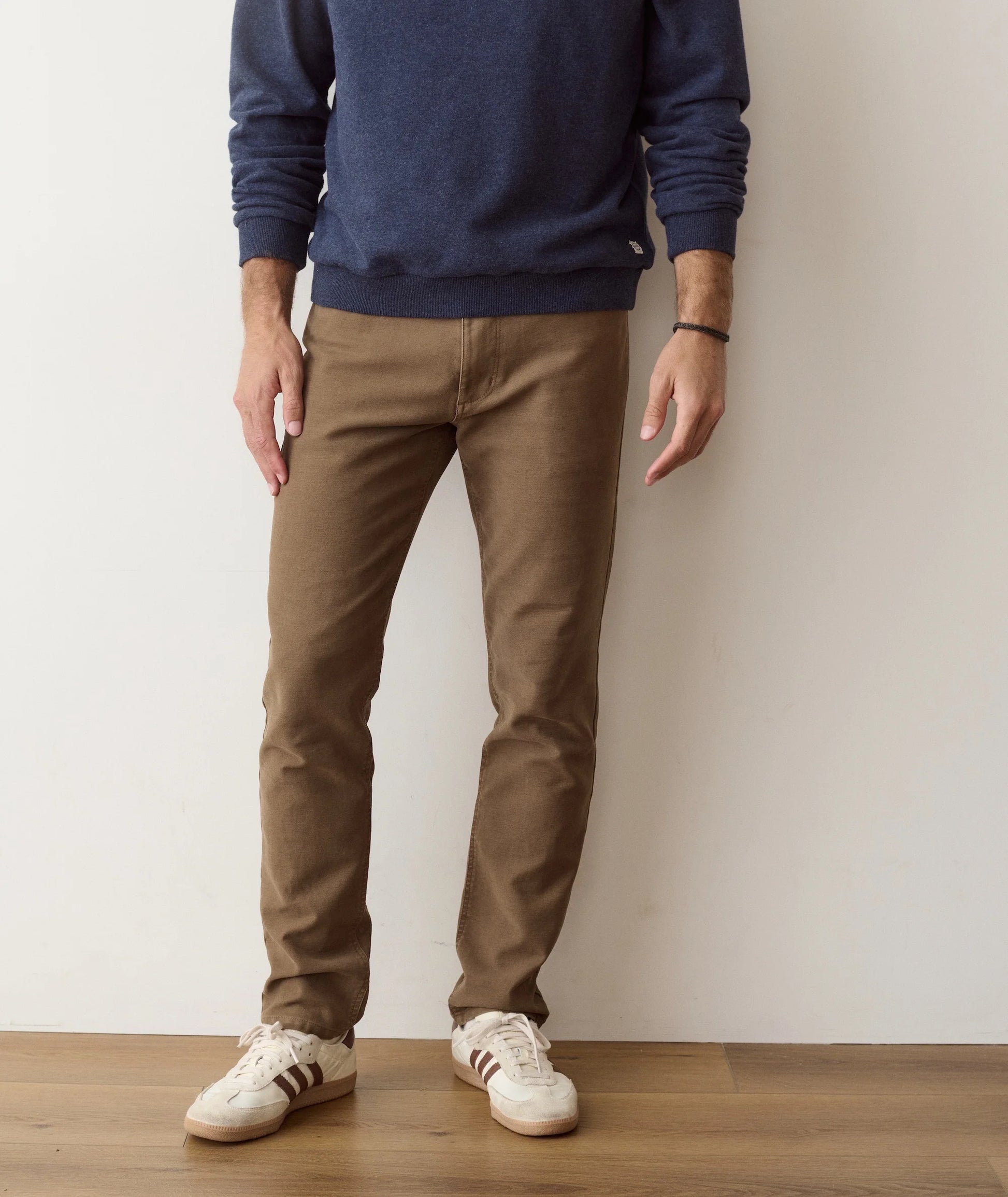 A person wearing slim brown pants, a navy blue long-sleeve top, and white low-top sneakers, standing against a plain off-white wall on a light wood floor.