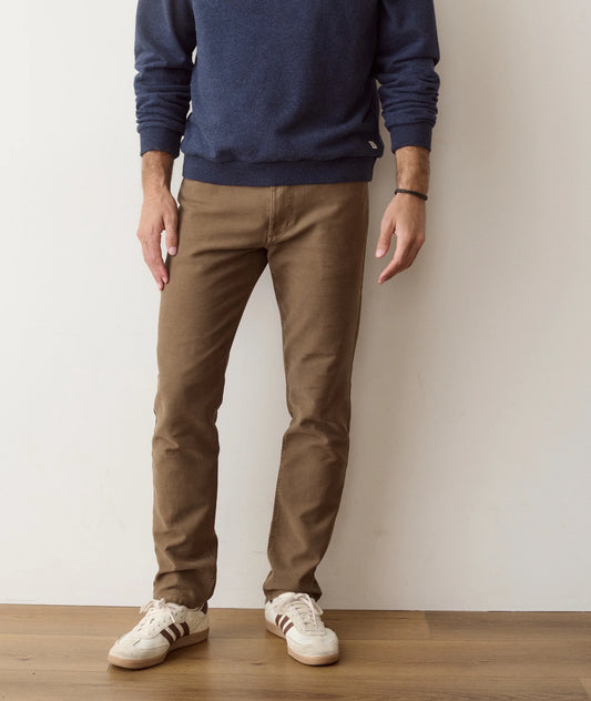 A person wearing slim brown pants, a navy blue long-sleeve top, and white low-top sneakers, standing against a plain off-white wall on a light wood floor.