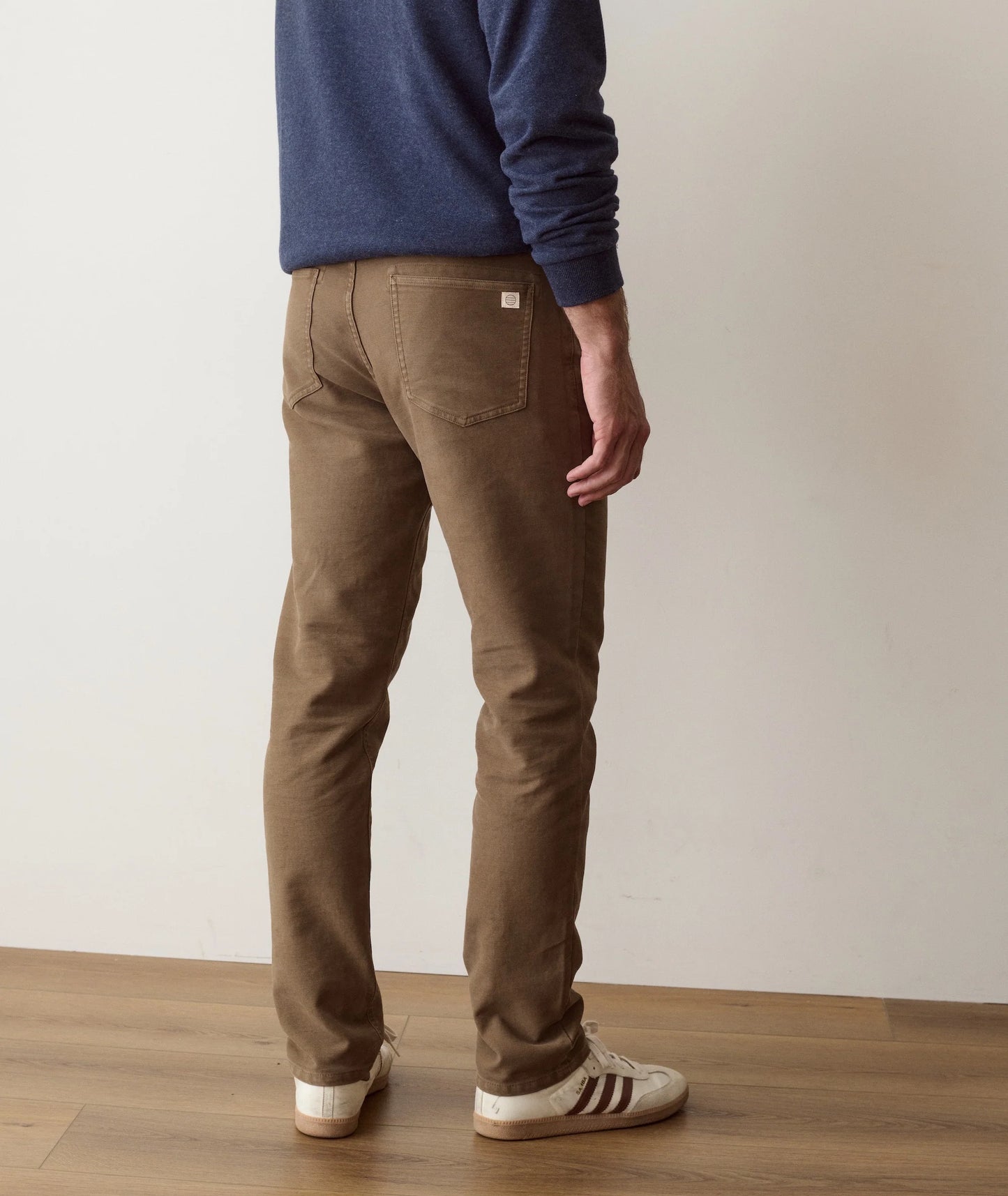 A person shown from the back wearing slim-fit brown pants, a navy blue long-sleeve top, and white sneakers with dark stripes, standing on a wooden floor against a plain off-white wall.