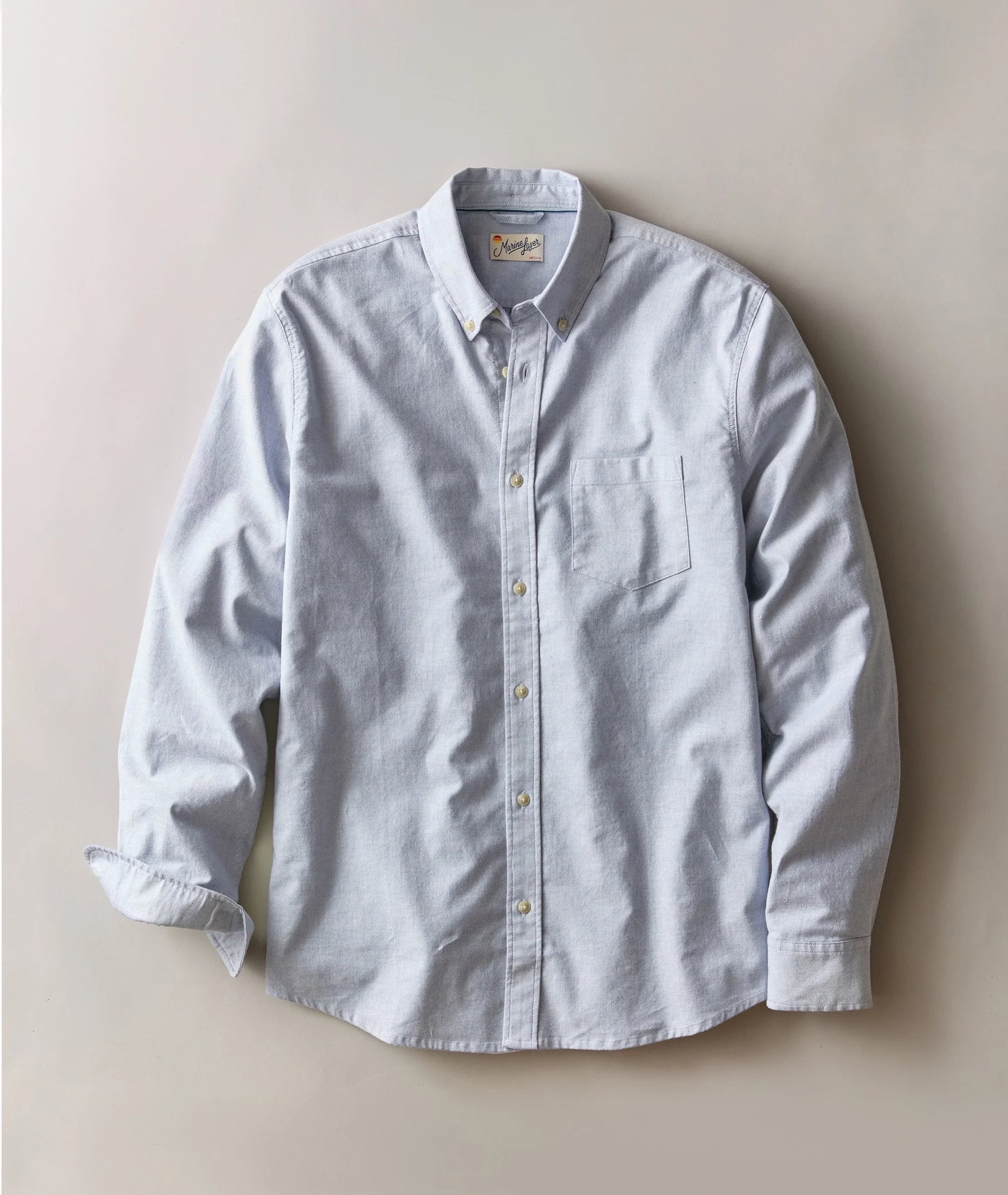 A light blue long-sleeve button-down shirt with a single chest pocket, laid flat against a neutral beige background.