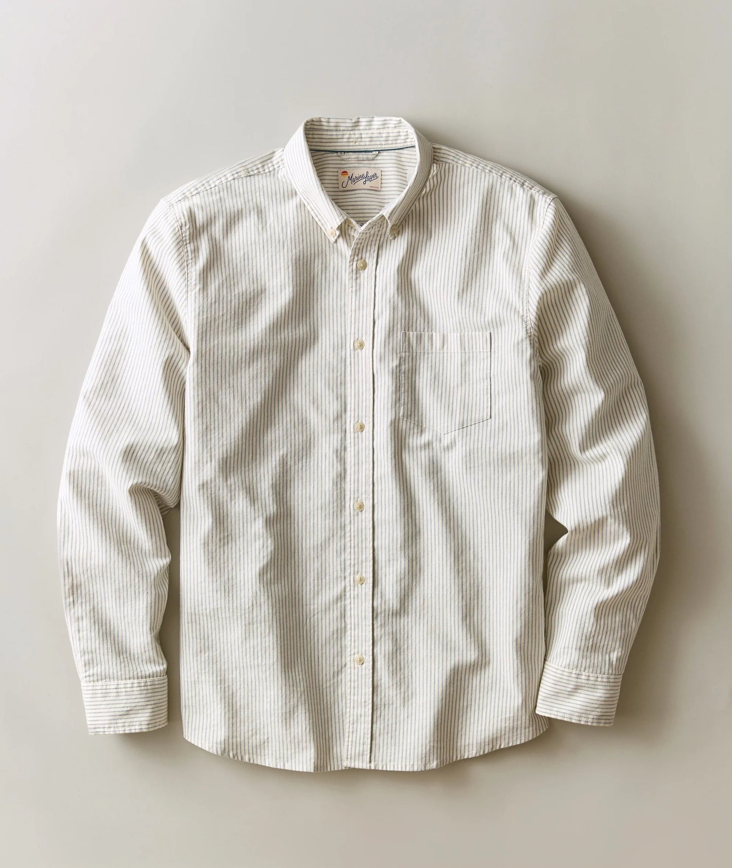 A long-sleeve button-down shirt in a cream and white micro-stripe pattern, laid flat against a neutral beige background.