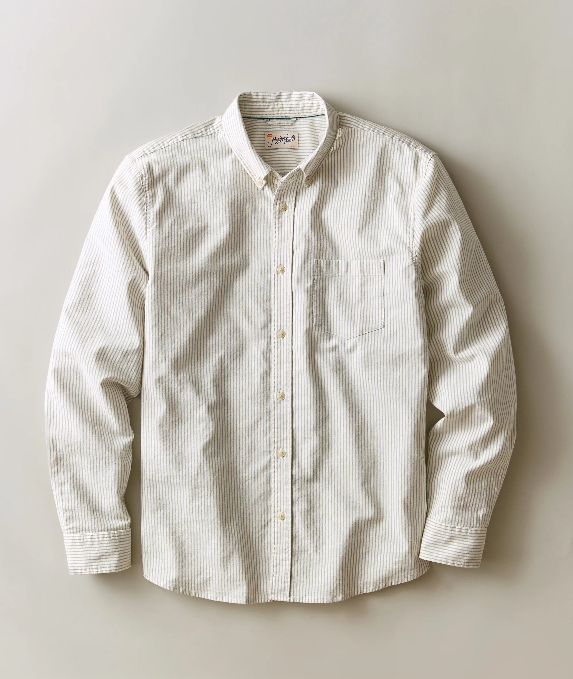 A long-sleeve button-down shirt in a cream and white micro-stripe pattern, laid flat against a neutral beige background.
