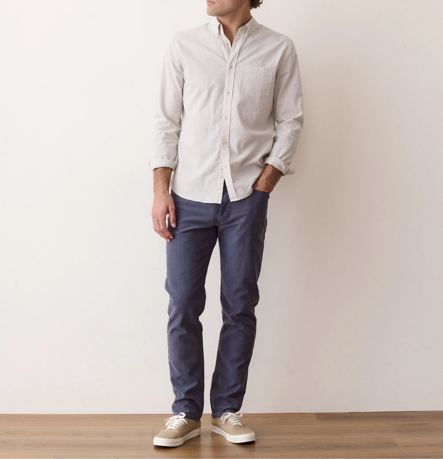 A man wearing a light beige long-sleeve button-down shirt, navy blue pants, and beige sneakers, standing against a plain off-white wall.