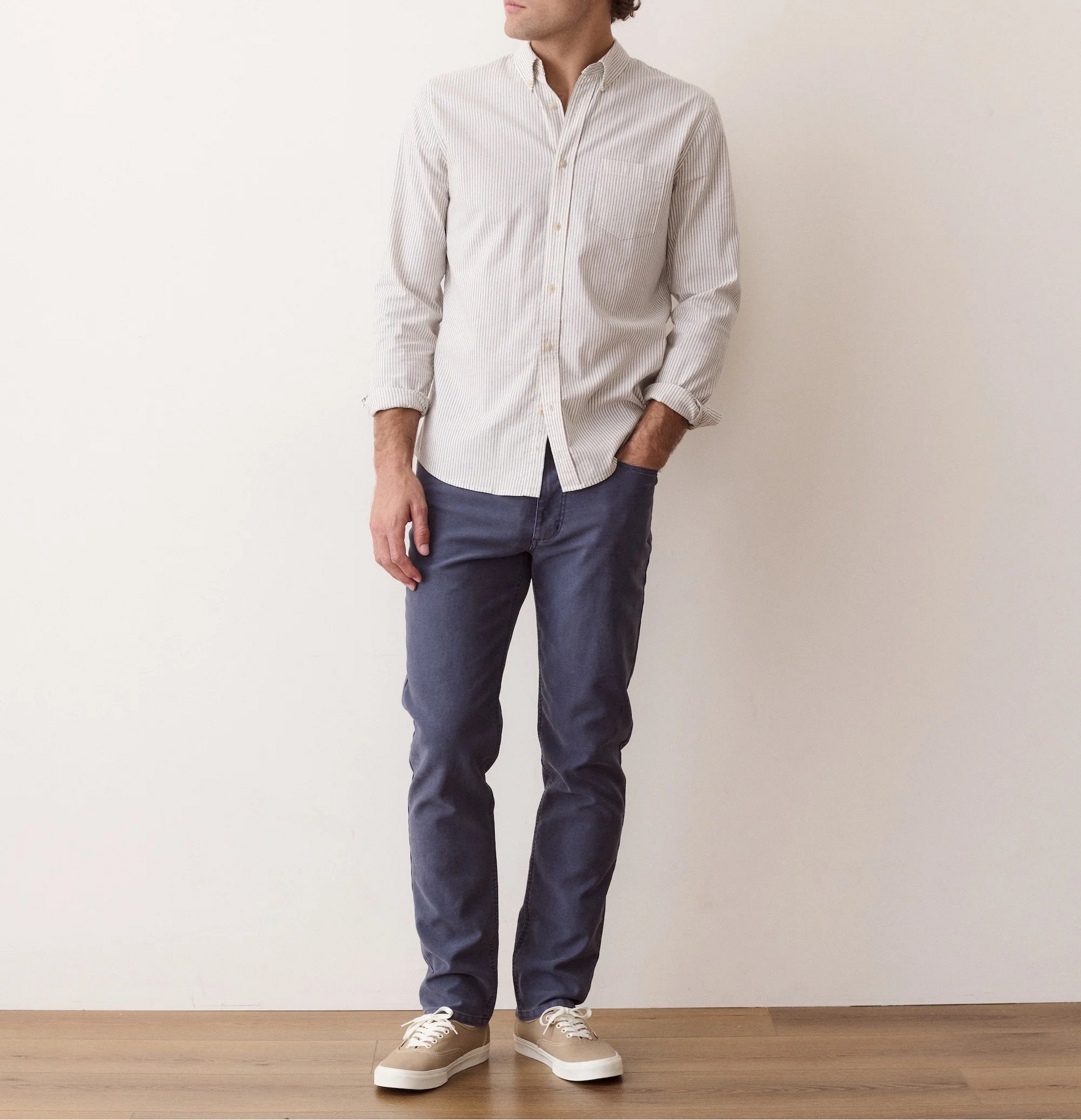 A man wearing a light beige long-sleeve button-down shirt, navy blue pants, and beige sneakers, standing against a plain off-white wall.