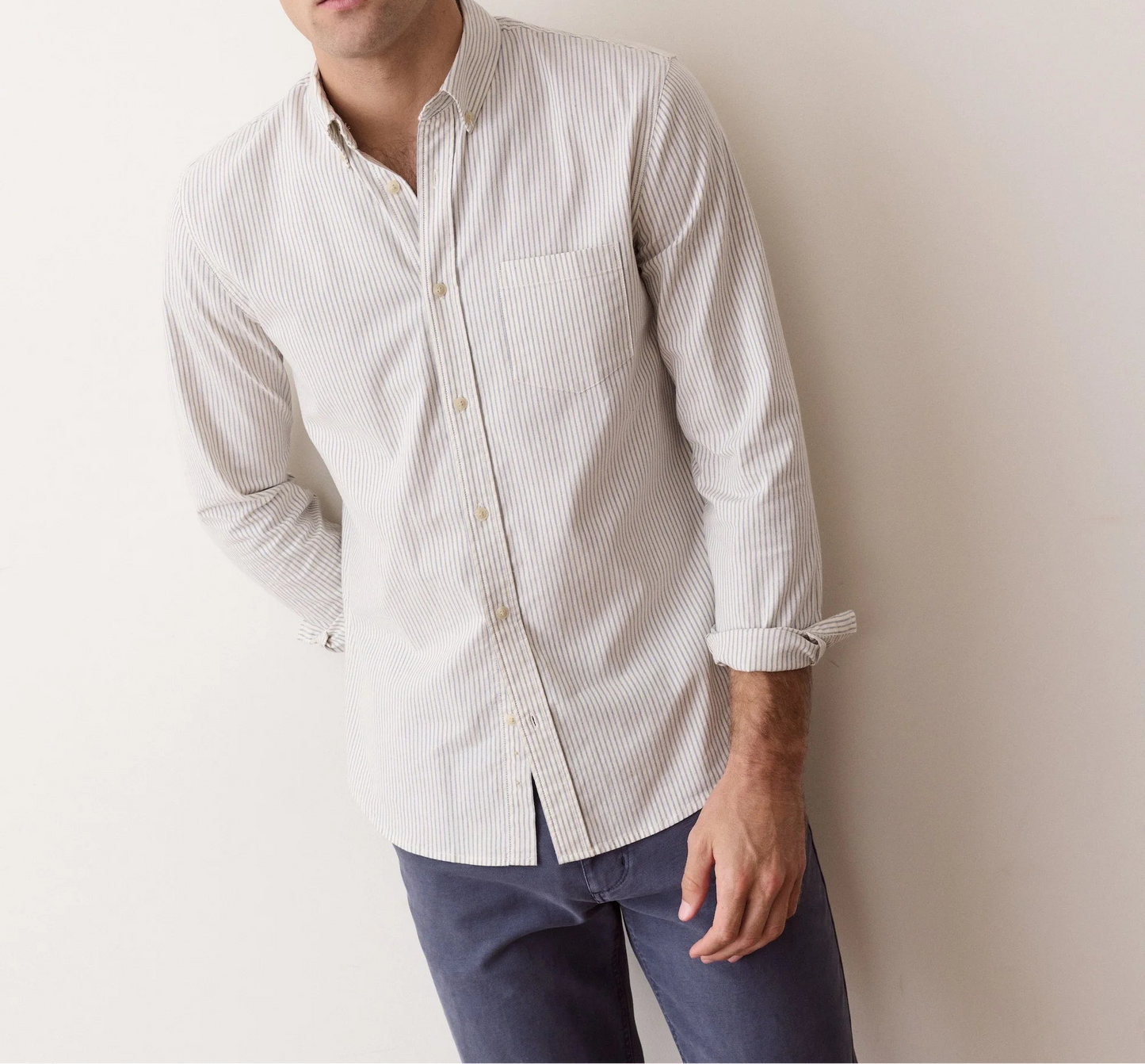 A man with curly brown hair wearing a light beige and white micro-striped long-sleeve button-down shirt and dark blue jeans, leaning against a plain off-white wall.