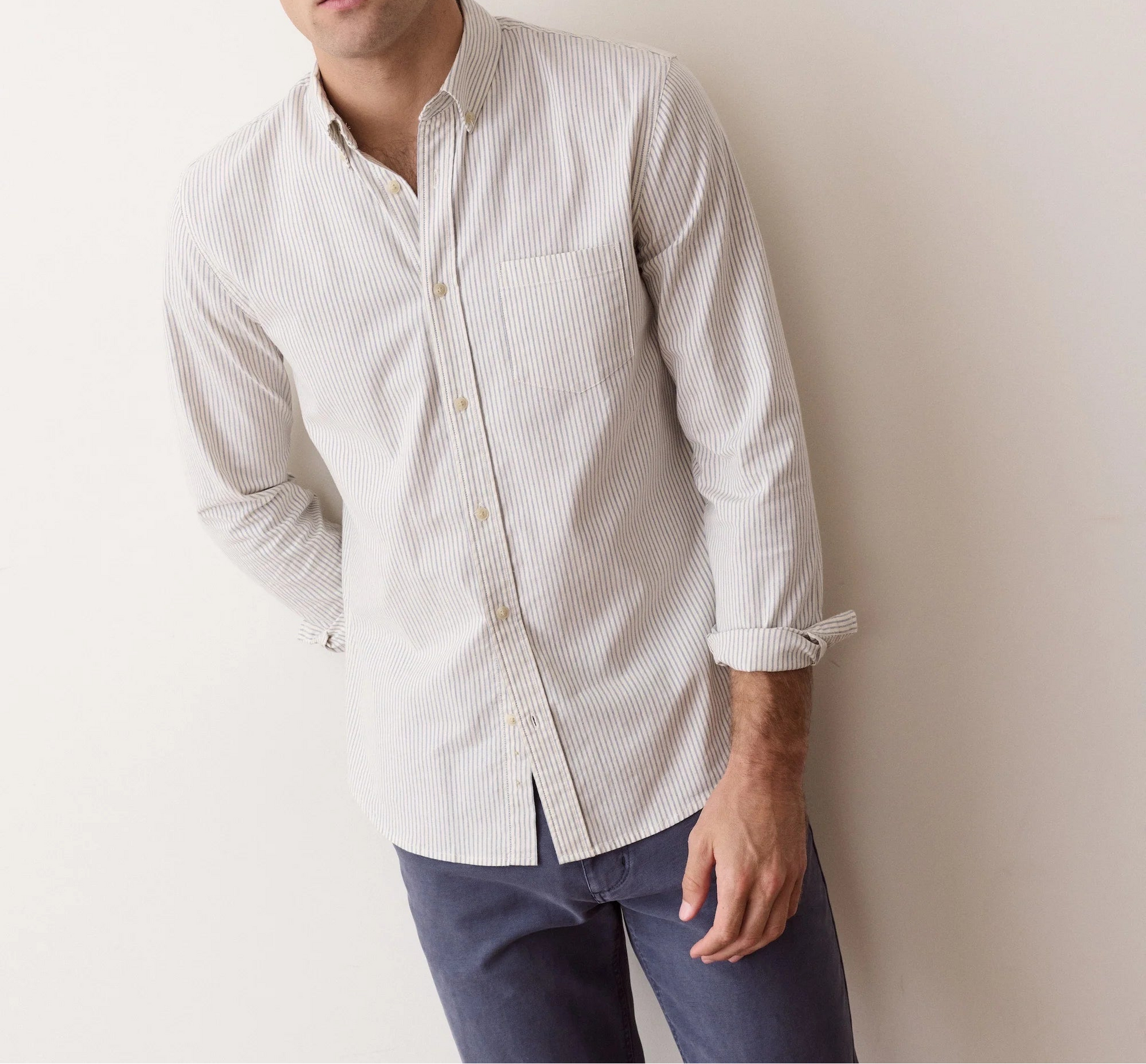 A man with curly brown hair wearing a light beige and white micro-striped long-sleeve button-down shirt and dark blue jeans, leaning against a plain off-white wall.