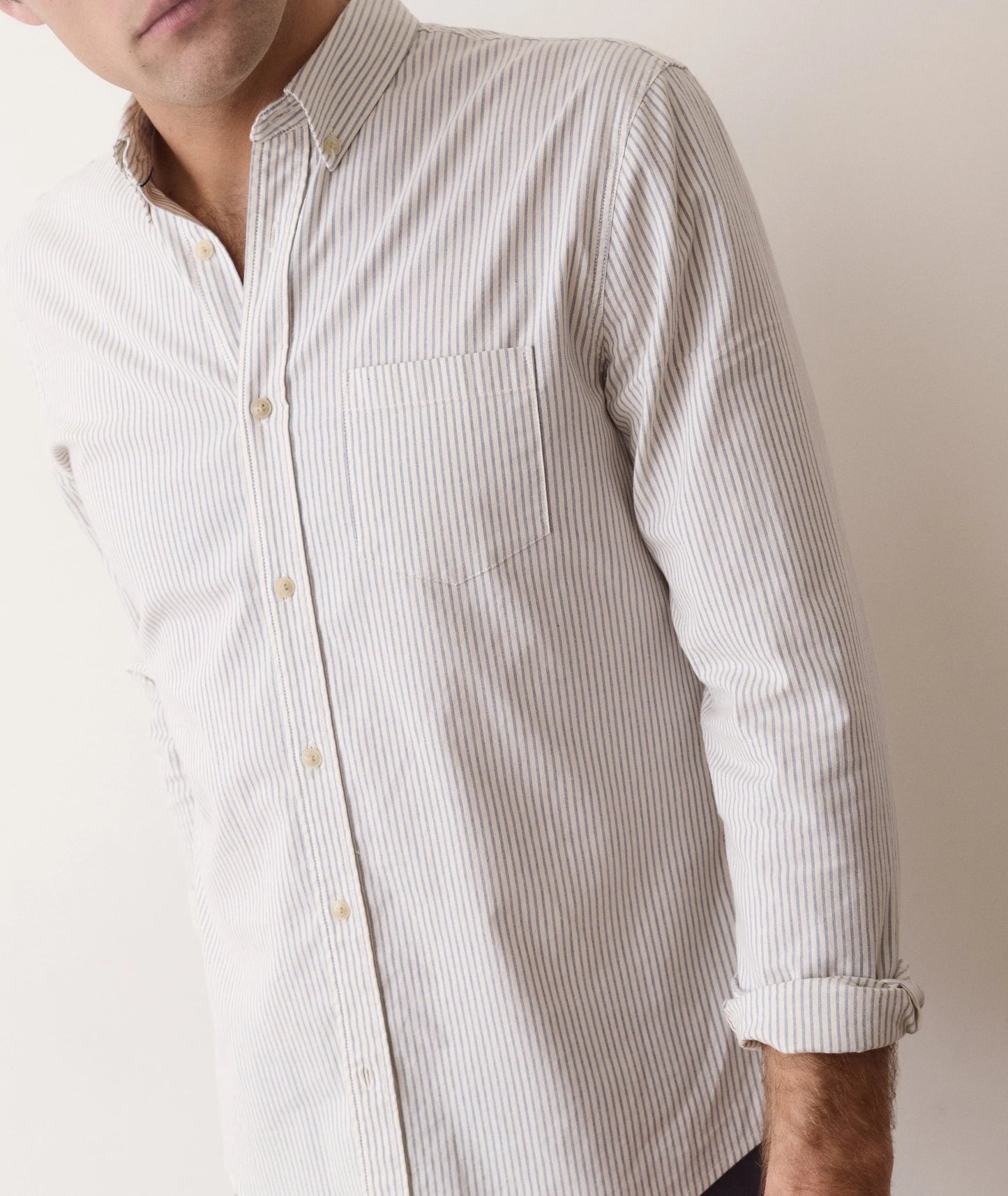 A close-up of a man wearing a long-sleeve button-down shirt with thin vertical stripes in off-white and beige, featuring a chest pocket and light-colored buttons.