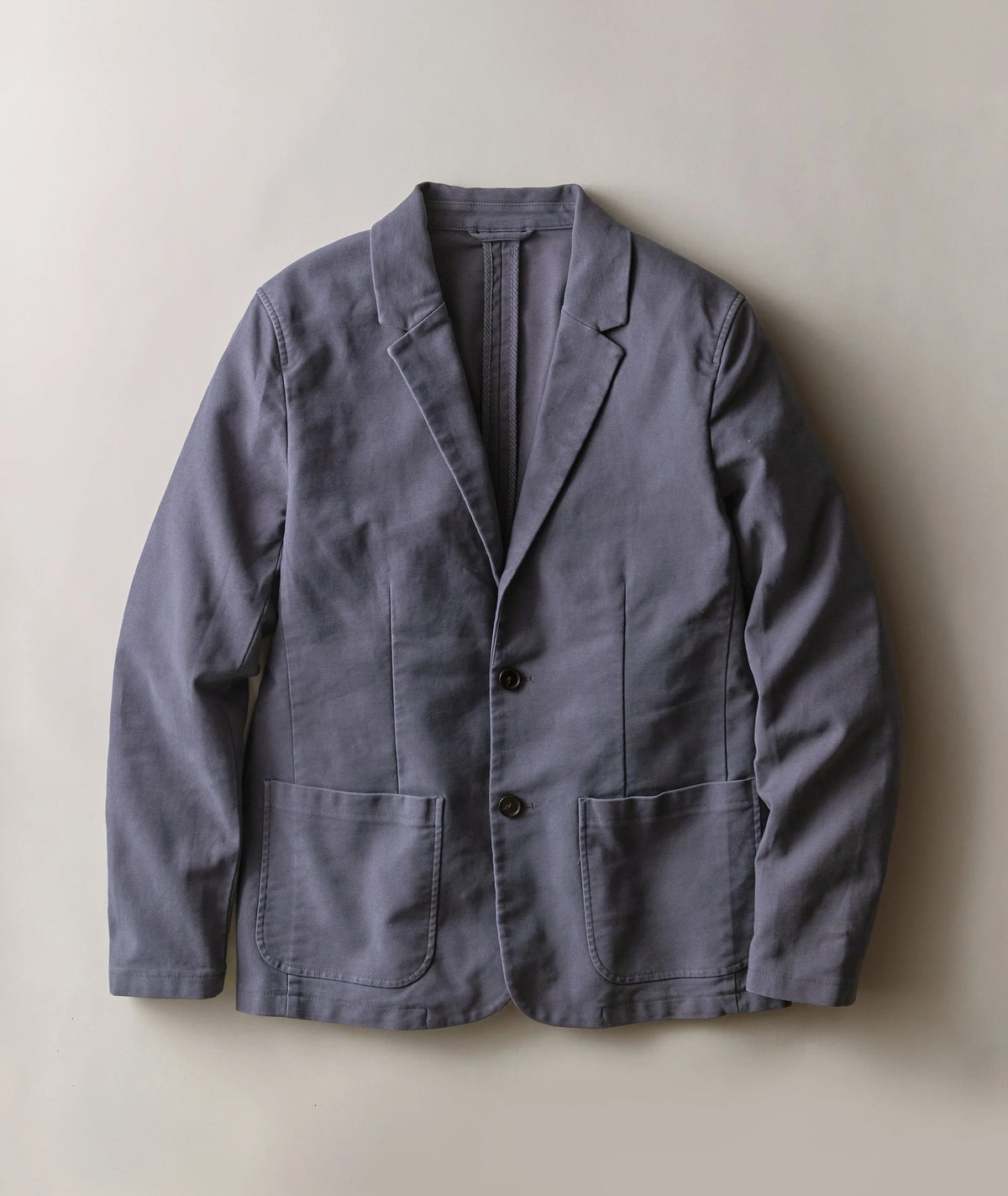 A slate blue-gray linen-blend blazer jacket with notch lapels, a two-button front closure, and two large front patch pockets, laid flat against a neutral beige background.