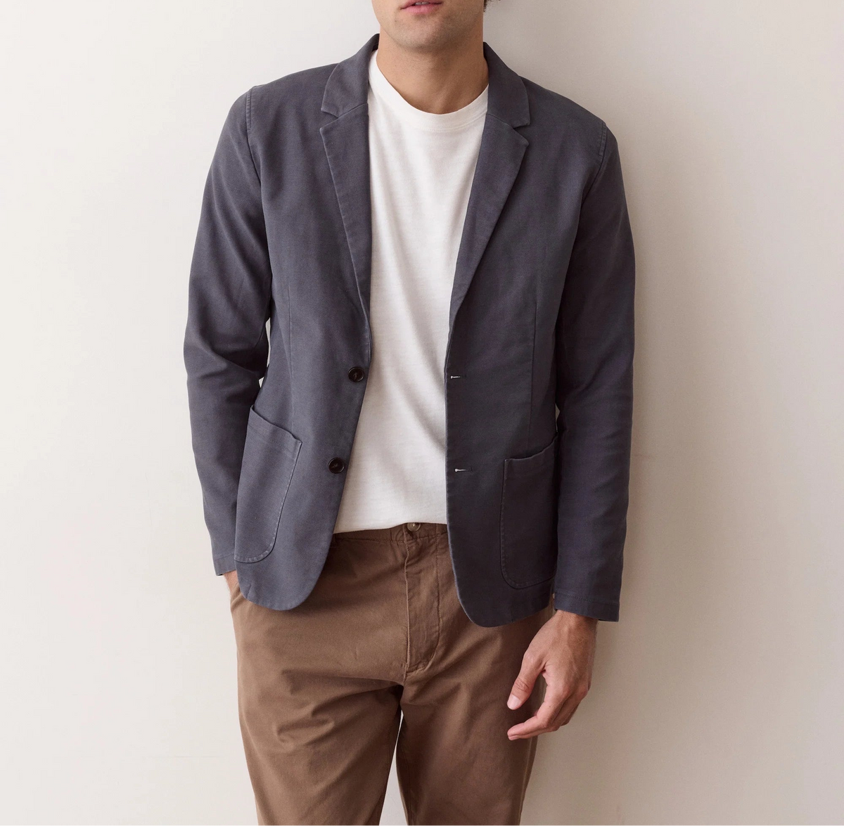 A man wearing a dark slate blue blazer over a white t-shirt and brown pants, standing against a plain off-white wall.