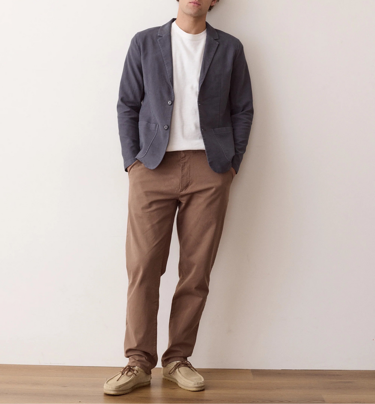 A man stands against a plain off-white wall wearing a dark slate blue blazer over a white t-shirt, brown trousers, and beige sneakers.