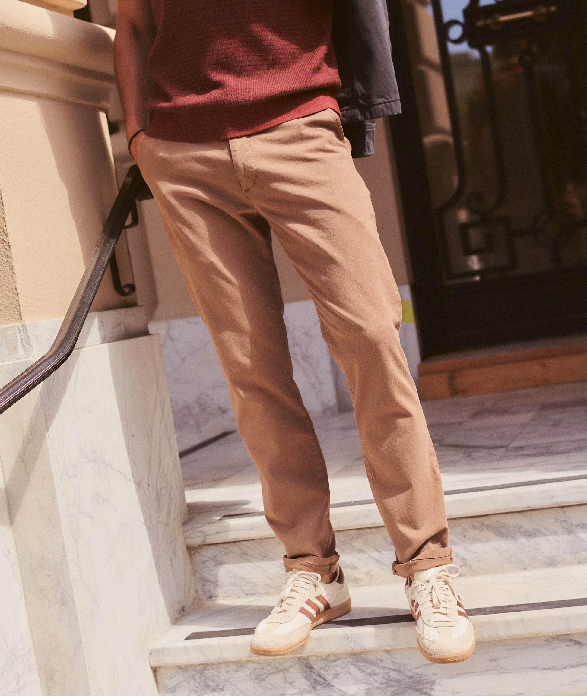 A person wearing slim-fit tan chinos, a dark red knit top, and cream sneakers with three stripes, standing on marble stairs.