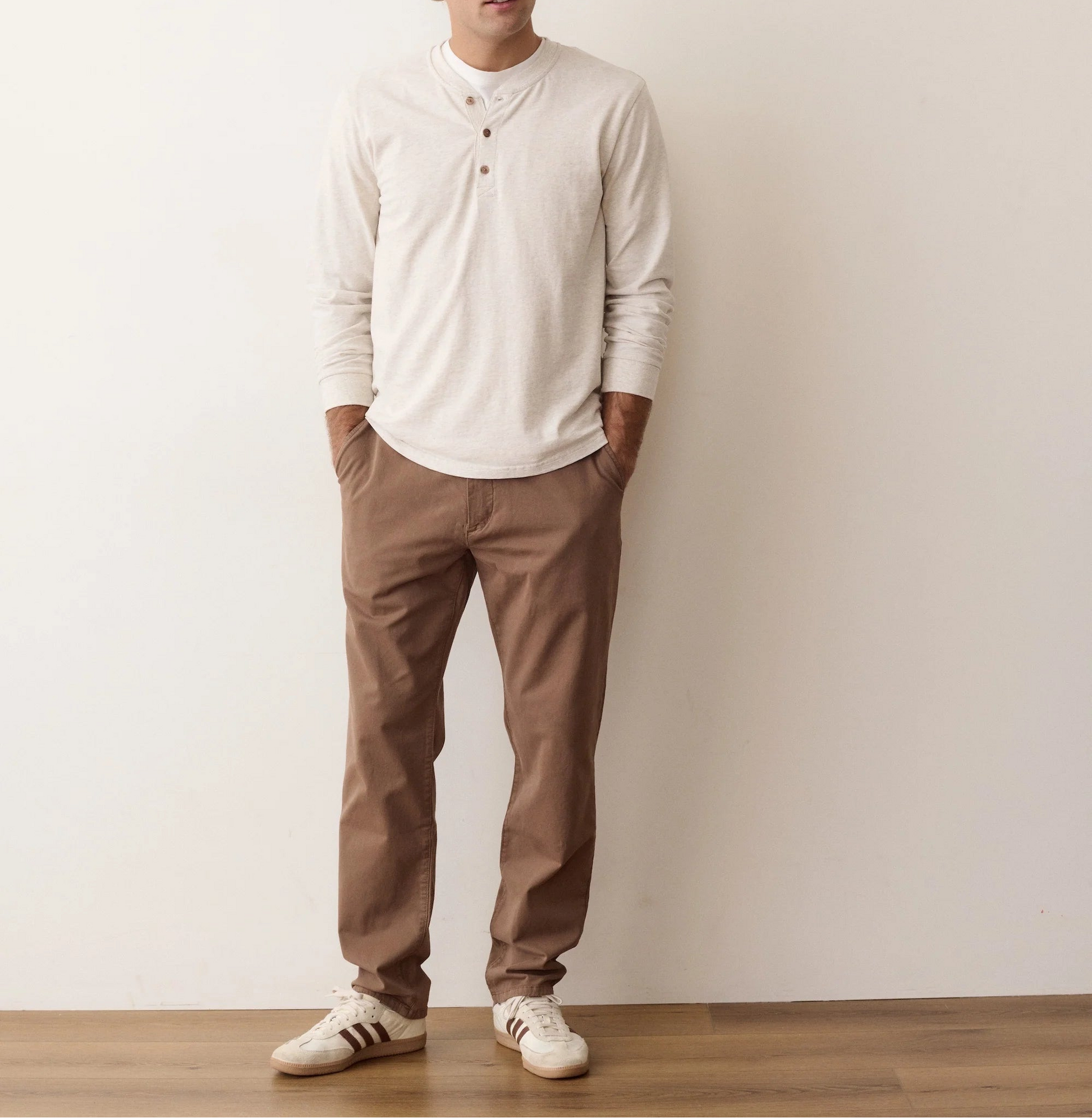 A man wearing a cream long-sleeve henley shirt, brown pants, and white sneakers stands against a plain off-white wall with a wooden floor.