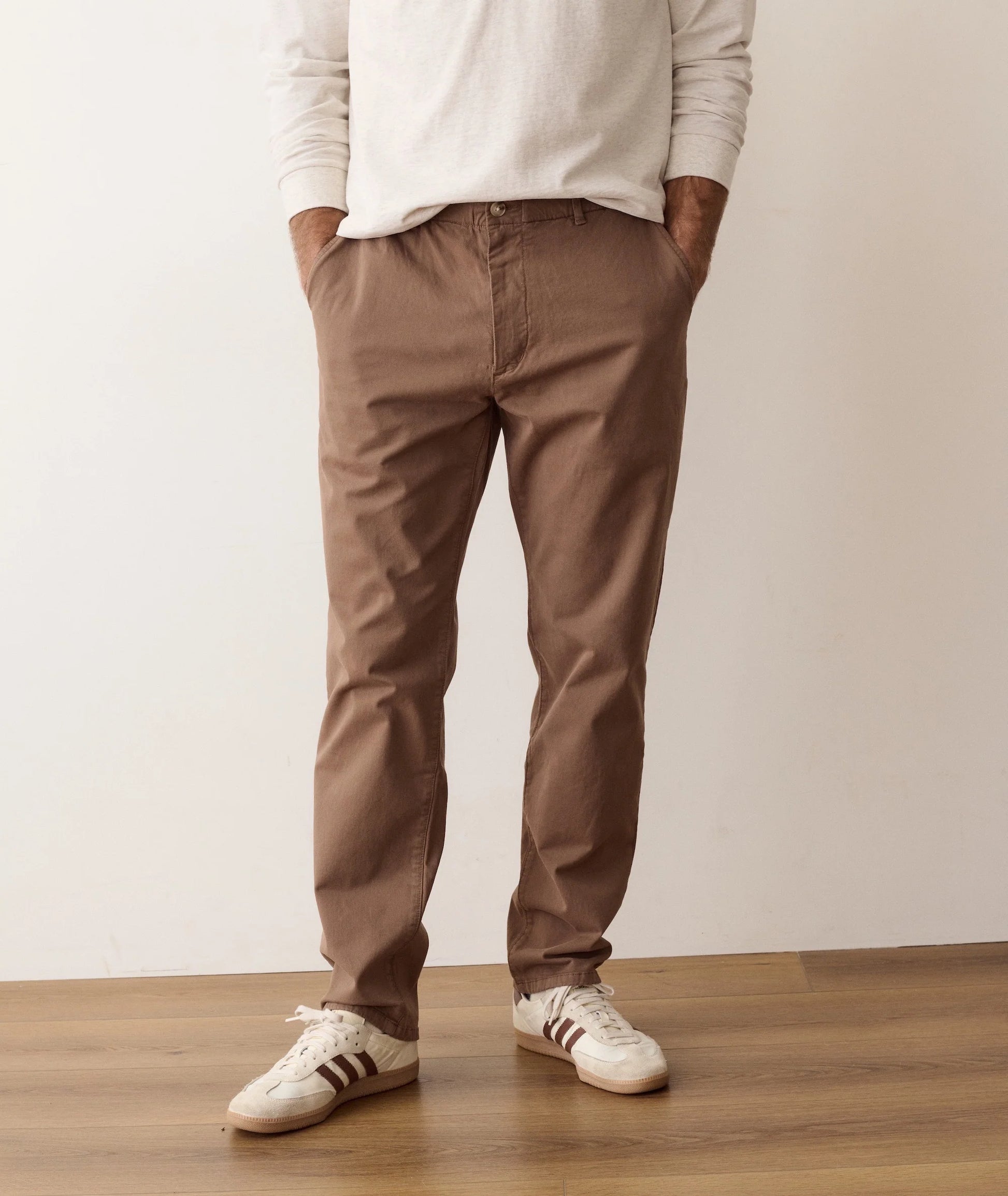 A person wearing slim-fit brown chino pants, a cream long-sleeve shirt, and white sneakers with dark stripes, standing on a wooden floor against a plain off-white wall.