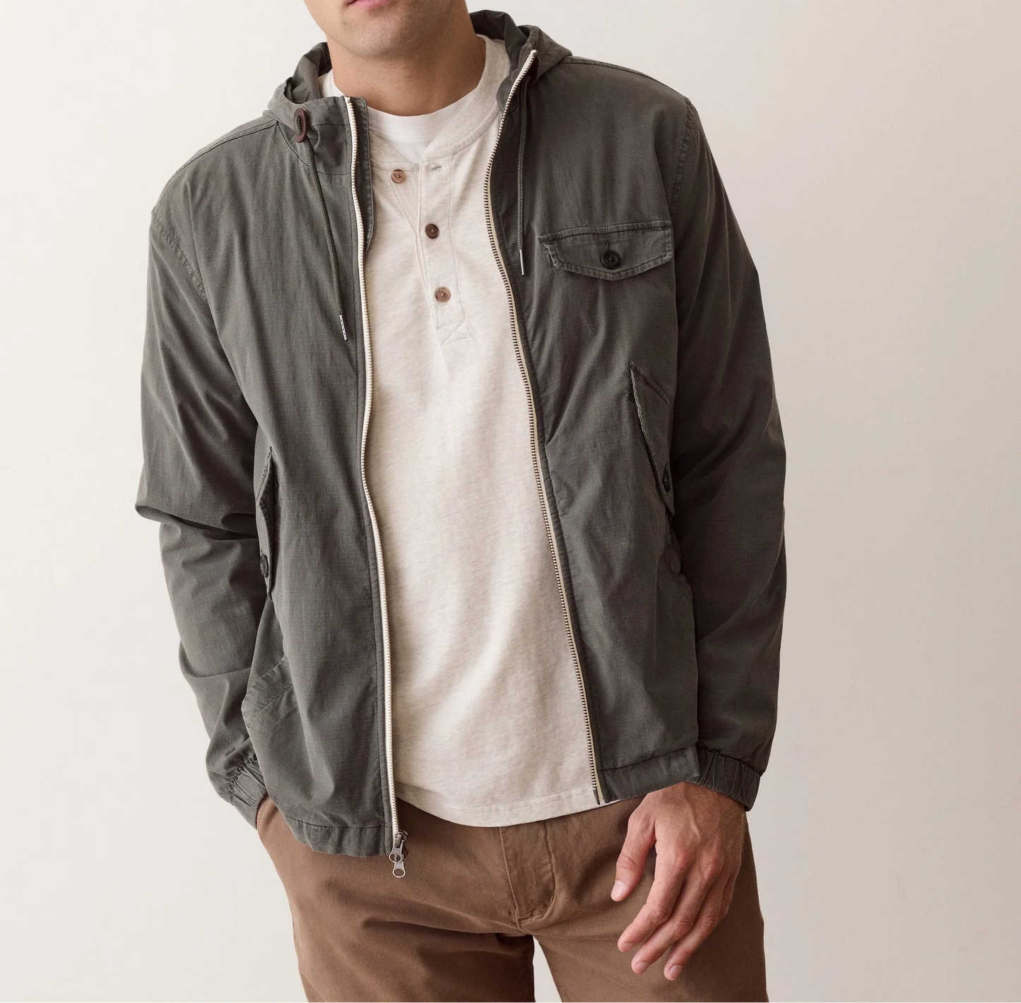 A man wearing a dark gray zip-up hooded jacket over a cream henley shirt and brown pants, photographed against a plain light beige studio background.