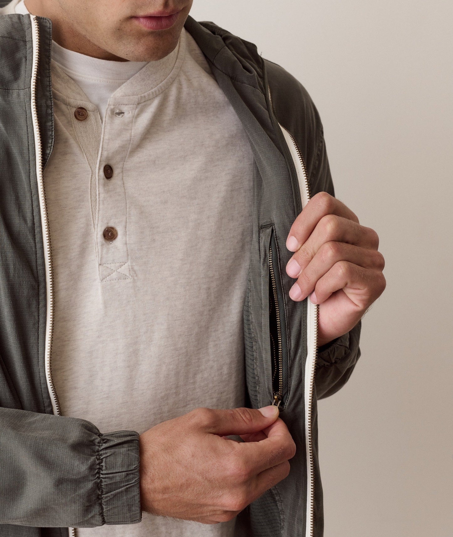 A close-up of a man wearing a dark gray zip-up jacket over a light beige henley shirt, with his hands unzipping the jacket.