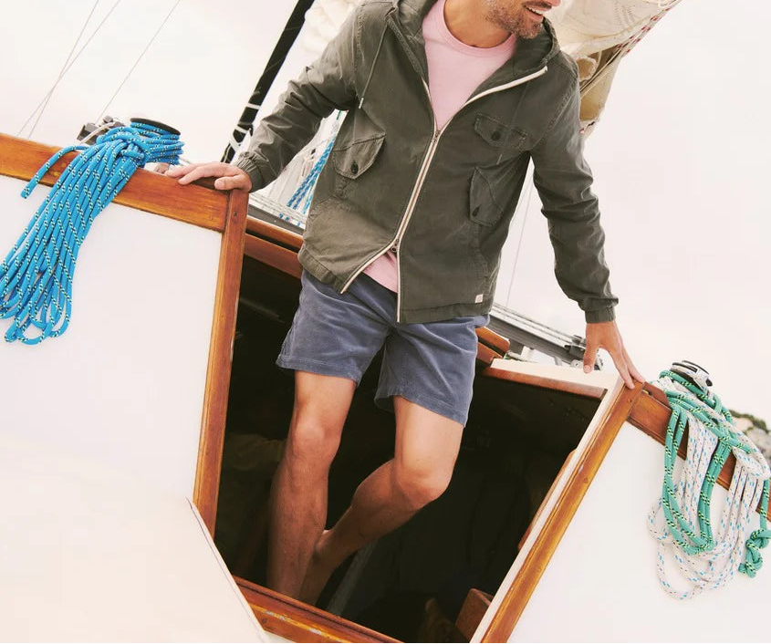 A man wearing a dark olive green zip-up hooded jacket, a light pink t-shirt, and blue shorts steps down into a wooden boat cabin. Blue and green ropes are visible on the deck against a bright white background.