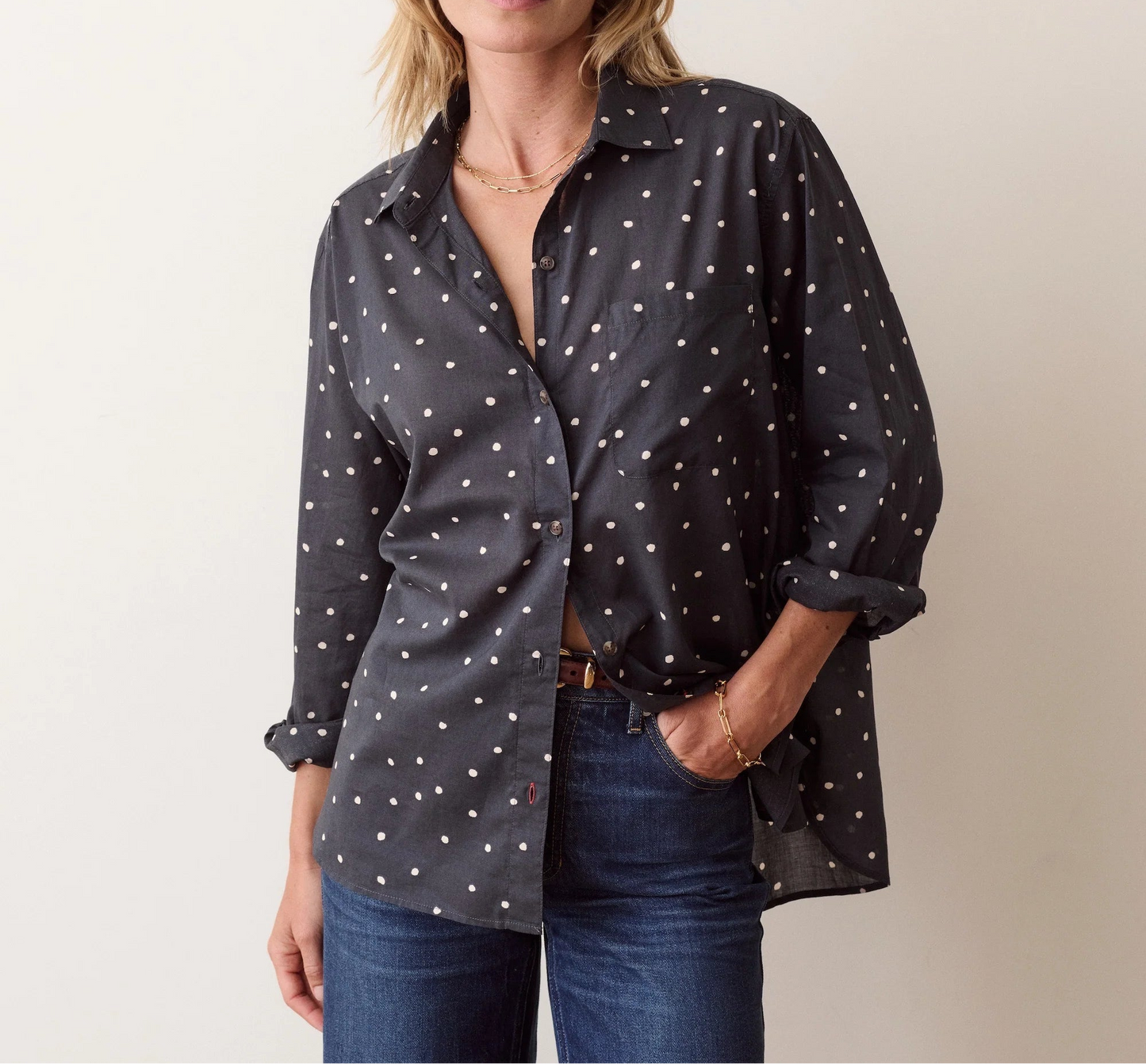 A woman modeling a dark gray button-up shirt with a white polka dot pattern, paired with blue jeans, against a plain off-white background.