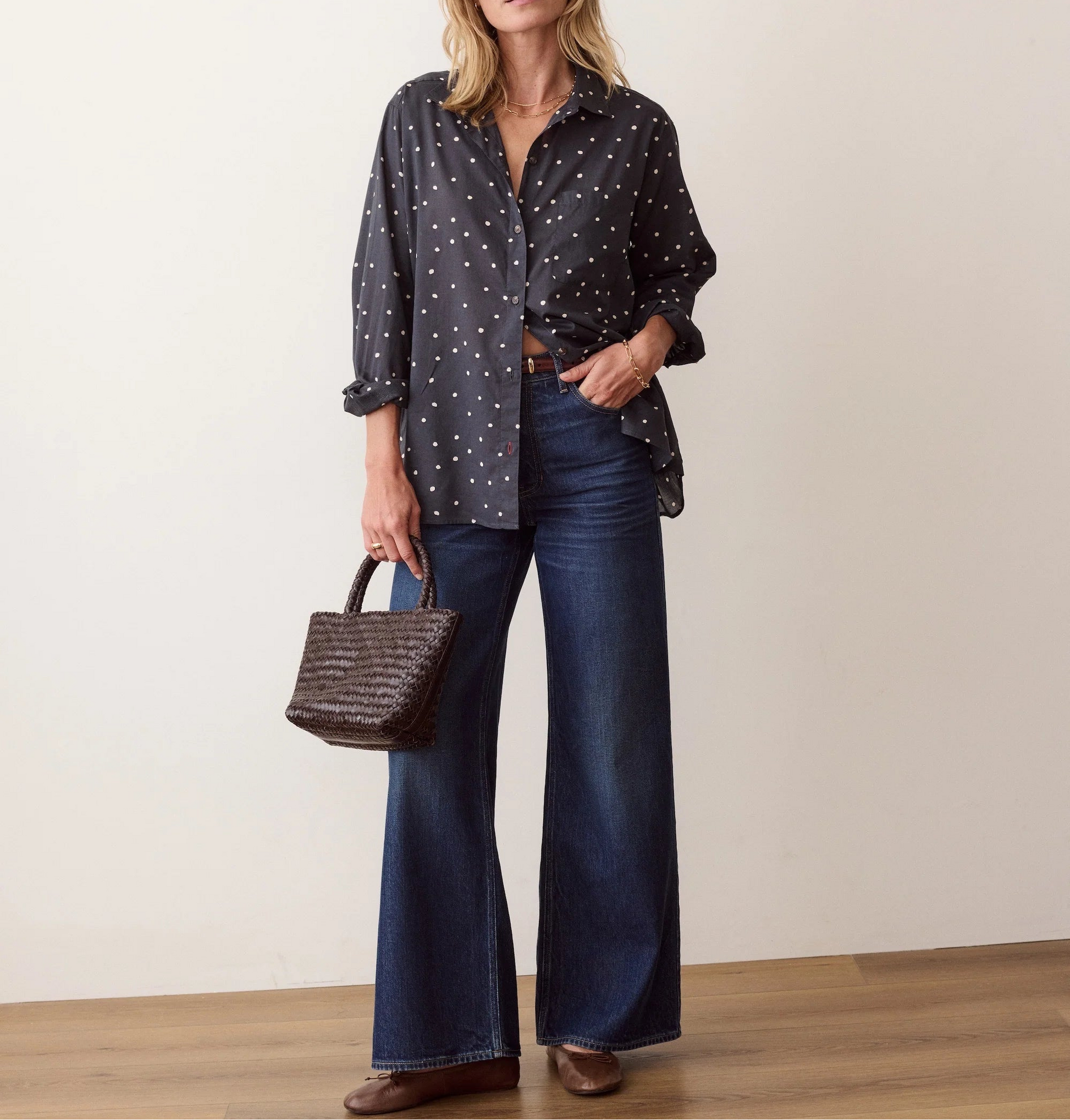 A woman stands in a studio wearing a dark navy button-up shirt with white polka dots, dark blue wide-leg jeans, brown shoes, and holding a brown woven handbag.
