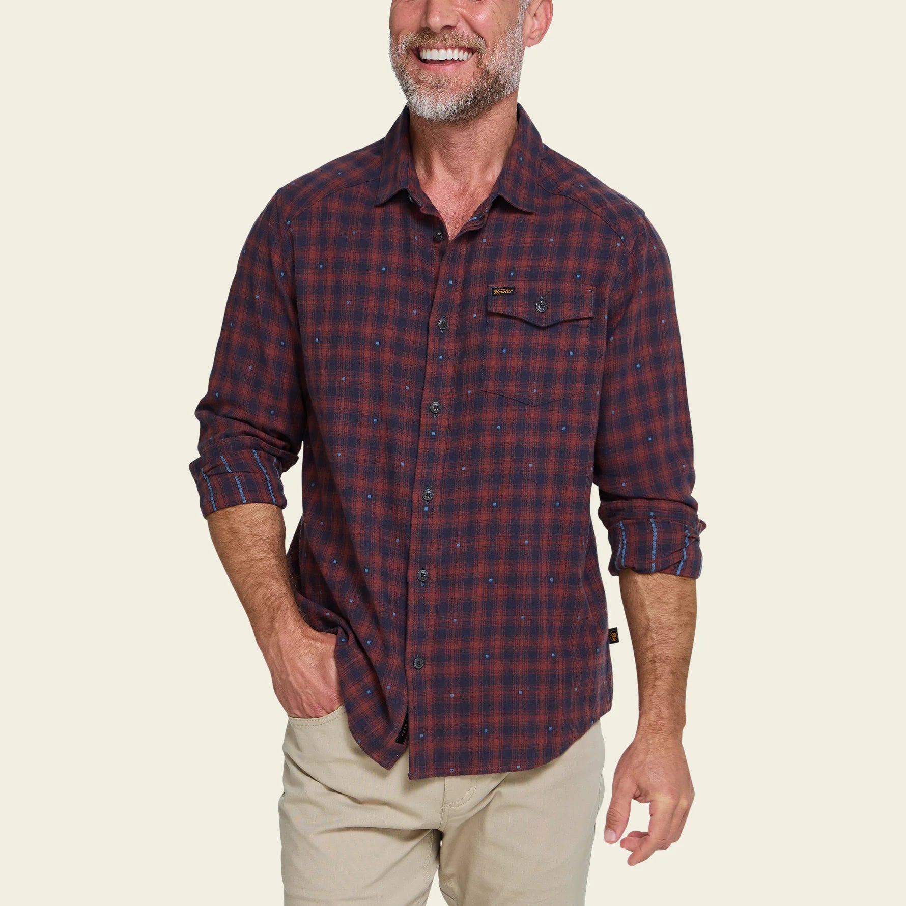 A smiling man wearing a long-sleeve button-up plaid shirt in dark red and navy, paired with khaki pants, against a plain white background.