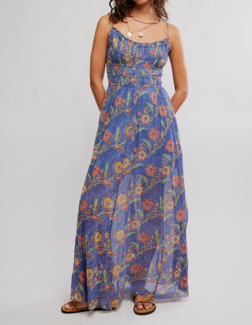 A woman wearing a sleeveless, floor-length blue maxi dress with a colorful floral print, paired with brown sandals and layered necklaces, standing against a light gray studio background.