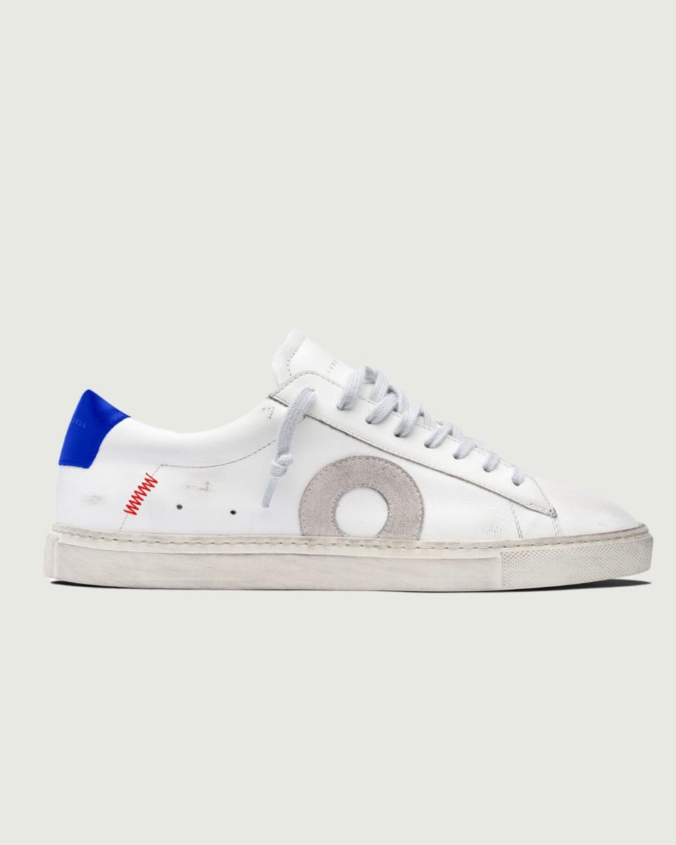 A white leather low-top sneaker with a large circular silver emblem on the side, white laces, a blue heel tab, and a distressed off-white sole, shown against a light gray background.