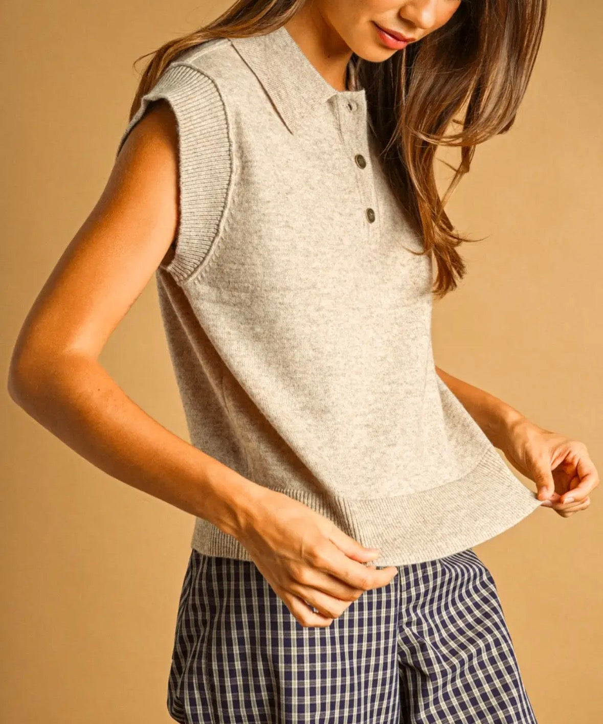 A woman wearing a dark gray sleeveless knit polo vest, posing against a solid warm brown studio background.