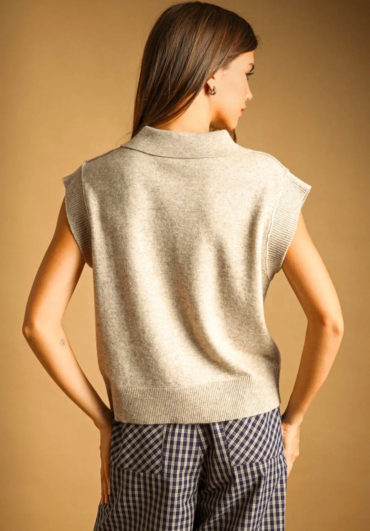 A woman shown from the back wearing a dark charcoal gray sleeveless knit sweater vest over a black and white checkered top, set against a solid warm tan background.