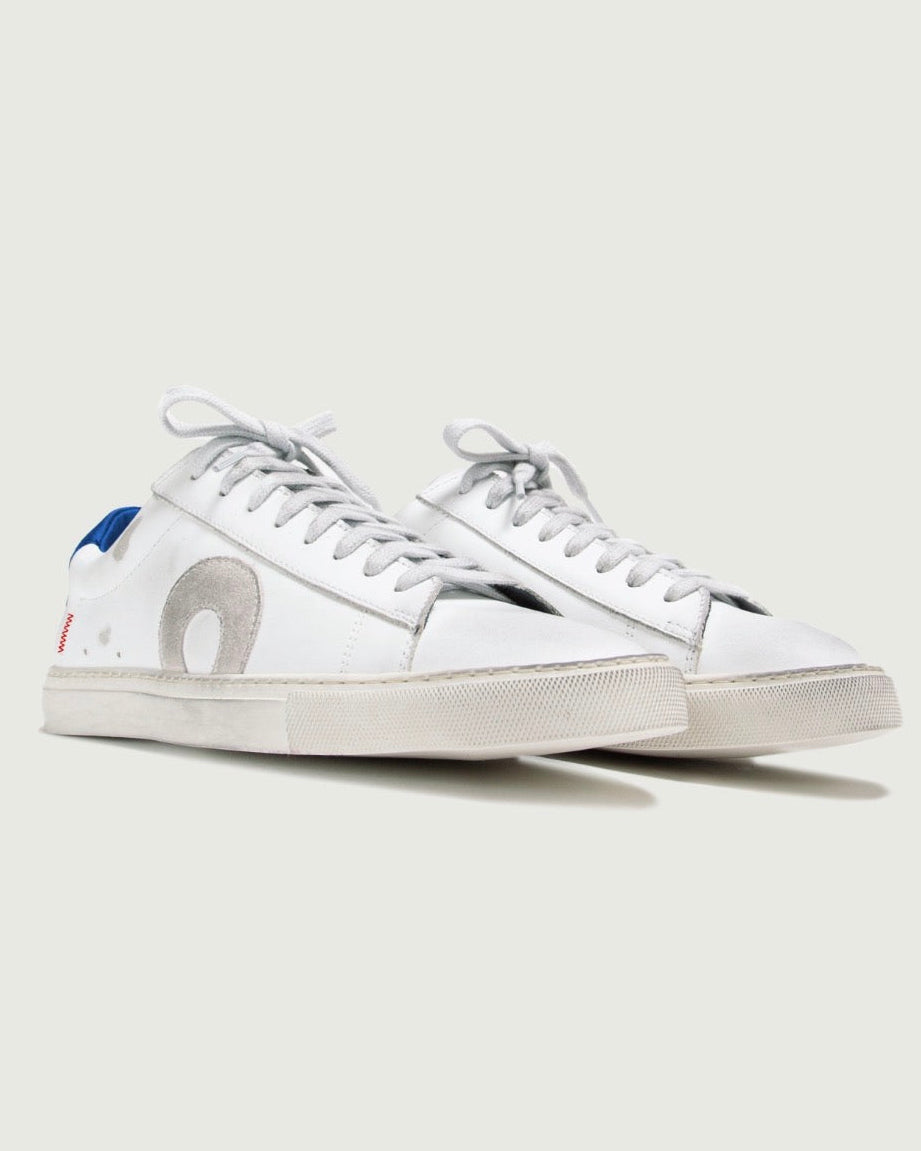 A pair of white leather low-top sneakers with white laces, featuring a large circular silver emblem on the side and a small blue and red accent on the heel tab, set against a solid light gray background.