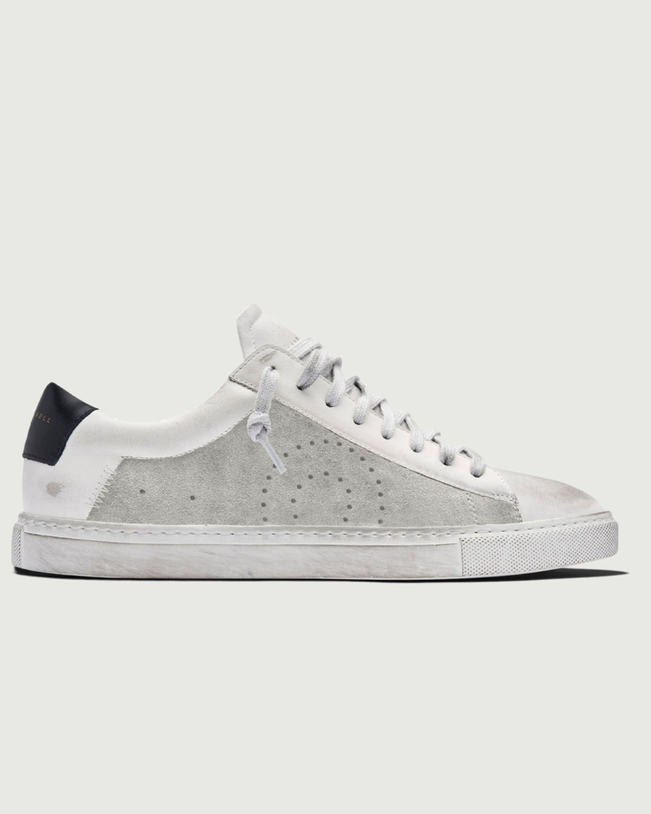 A low-top sneaker in white and light gray suede, featuring a perforated circular design on the side, white laces, and a black heel tab, shown against a light gray background.