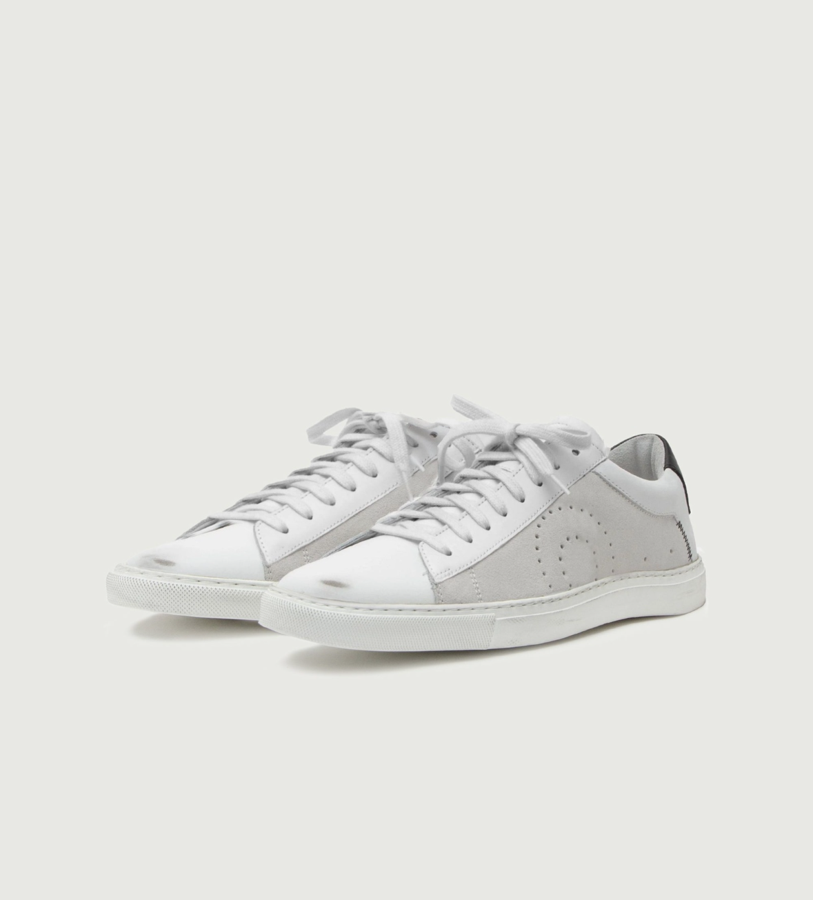 A pair of low-top sneakers in white leather and light gray suede, featuring perforated side details, white laces, and white soles, set against a solid off-white background.