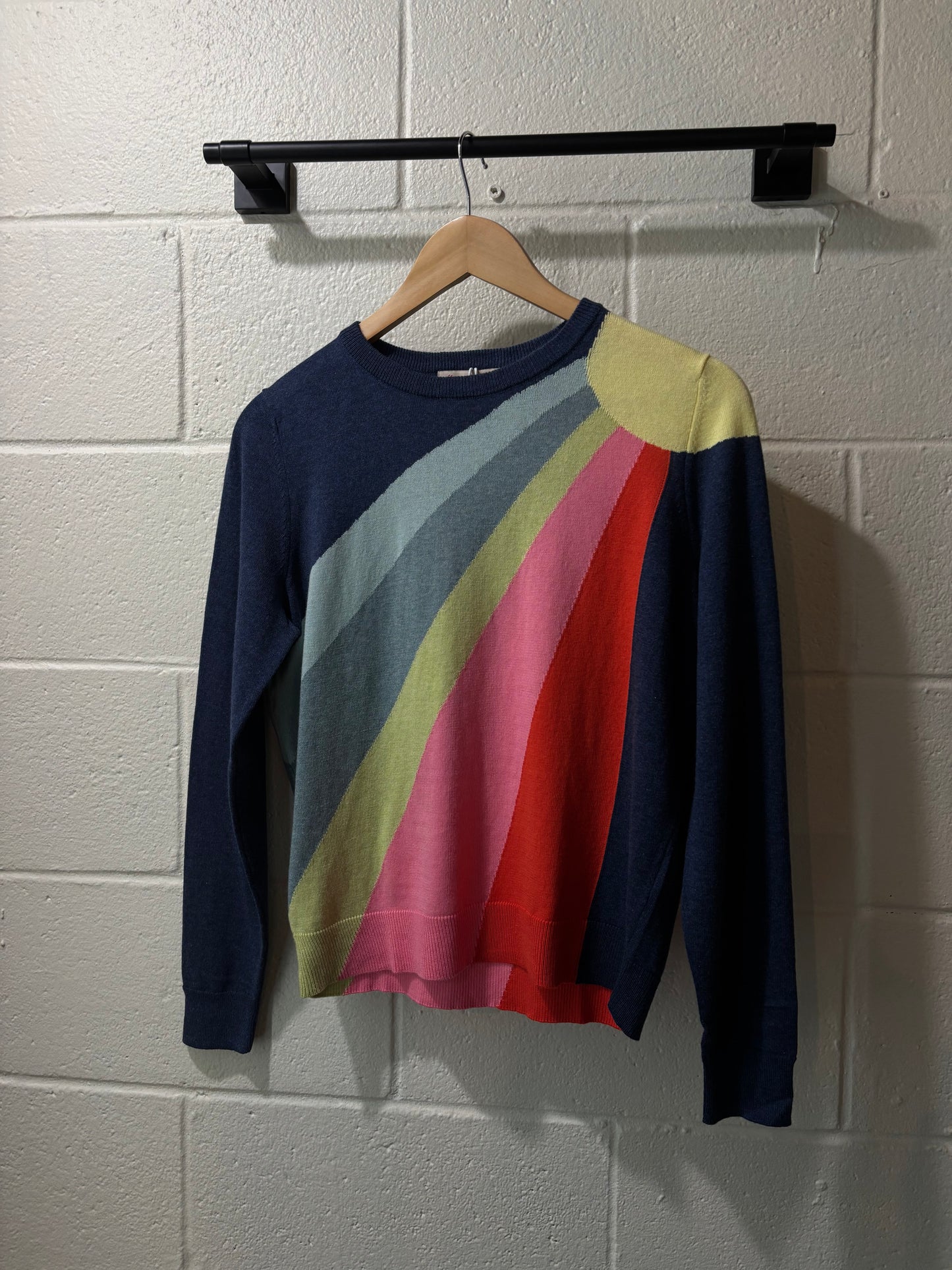 Colorful sweater with a geometric pattern hanging on a wooden hanger against a white brick wall.