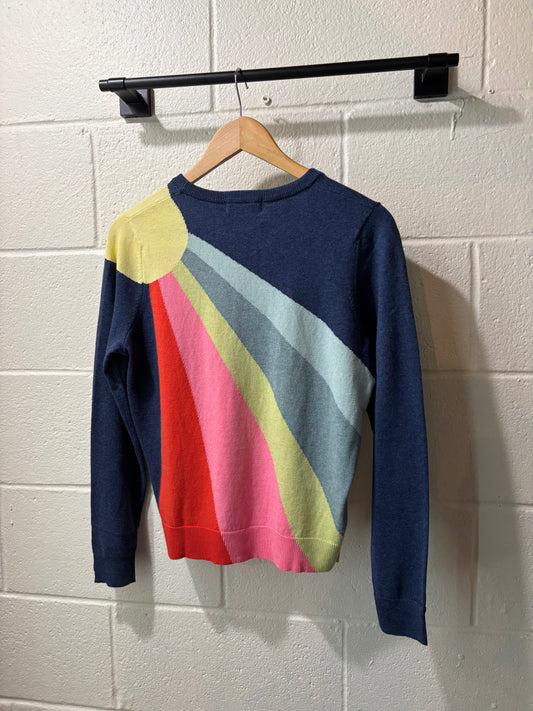 Colorful sweater with a rainbow pattern hanging on a wooden hanger against a white brick wall.
