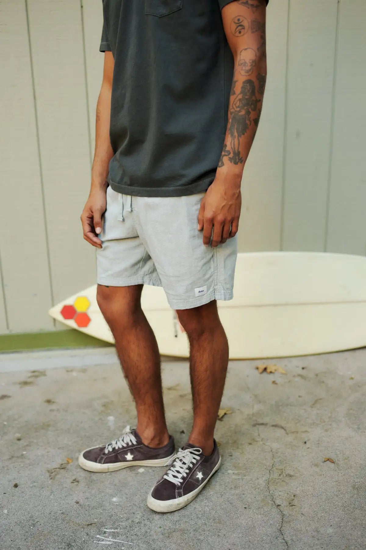 Person wearing a black shirt, gray shorts, and brown sneakers standing on a concrete surface with a surfboard in the background.