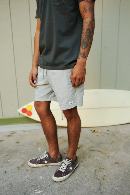 Person wearing a black shirt, gray shorts, and brown sneakers standing on a concrete surface with a surfboard in the background.