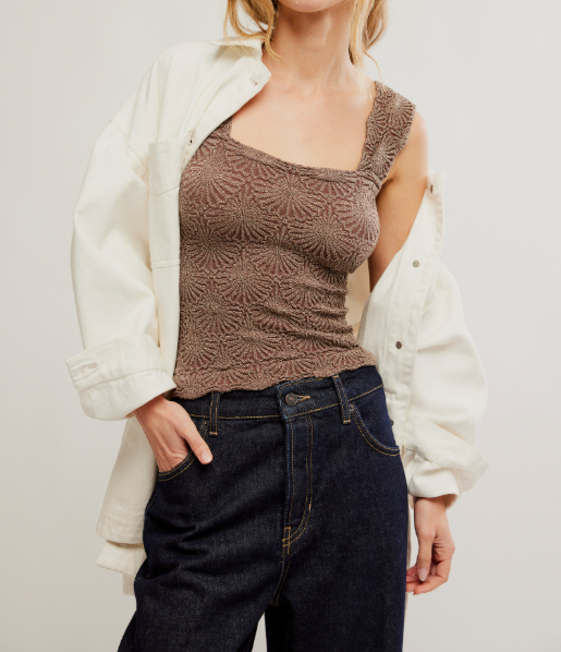 A woman wearing a brown textured knit tank top with a square neckline, styled under an oversized cream-colored denim shirt and paired with dark blue jeans, against a plain light gray background.