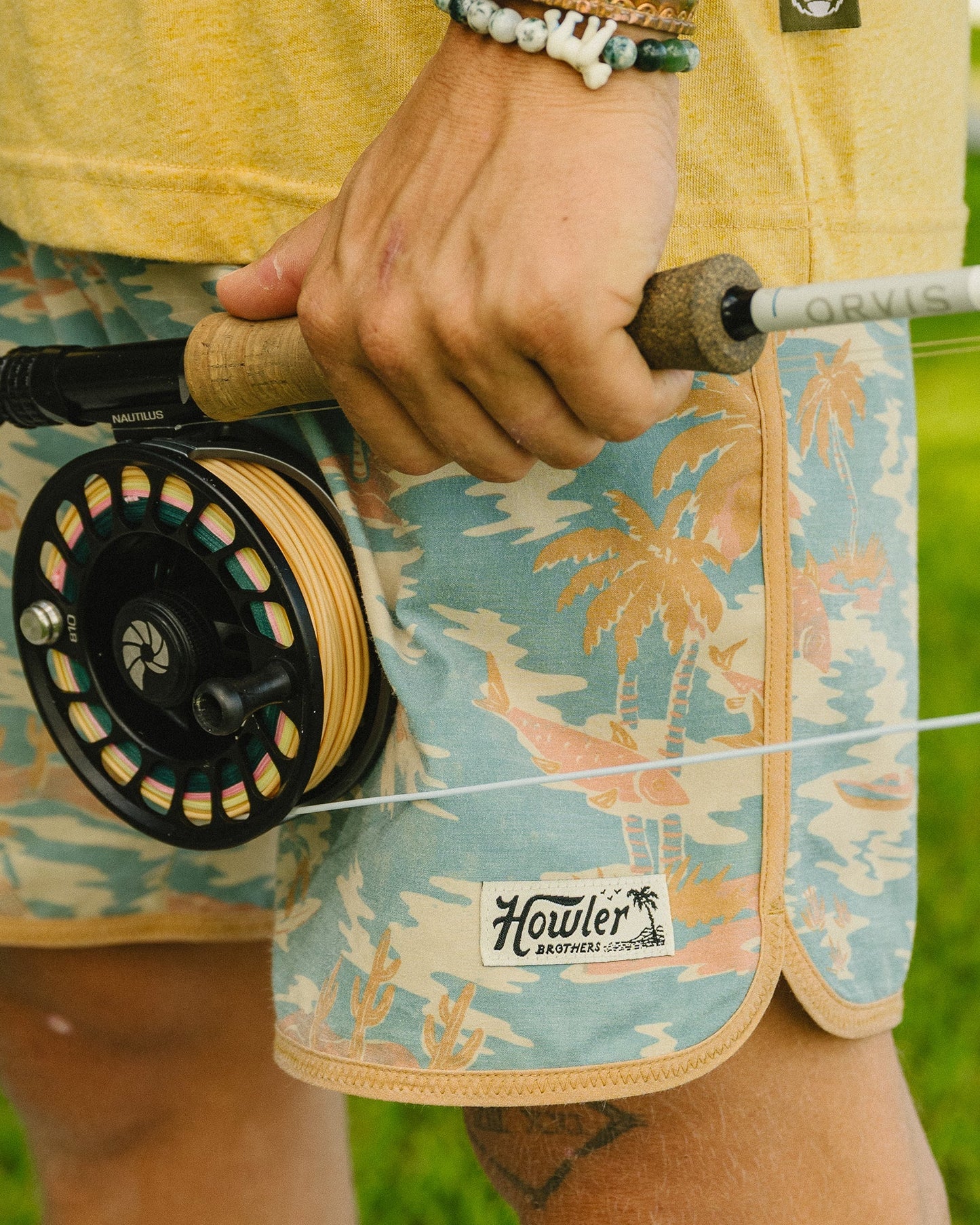 Person holding a fly fishing reel with 'Howler' branding on shorts, outdoors.