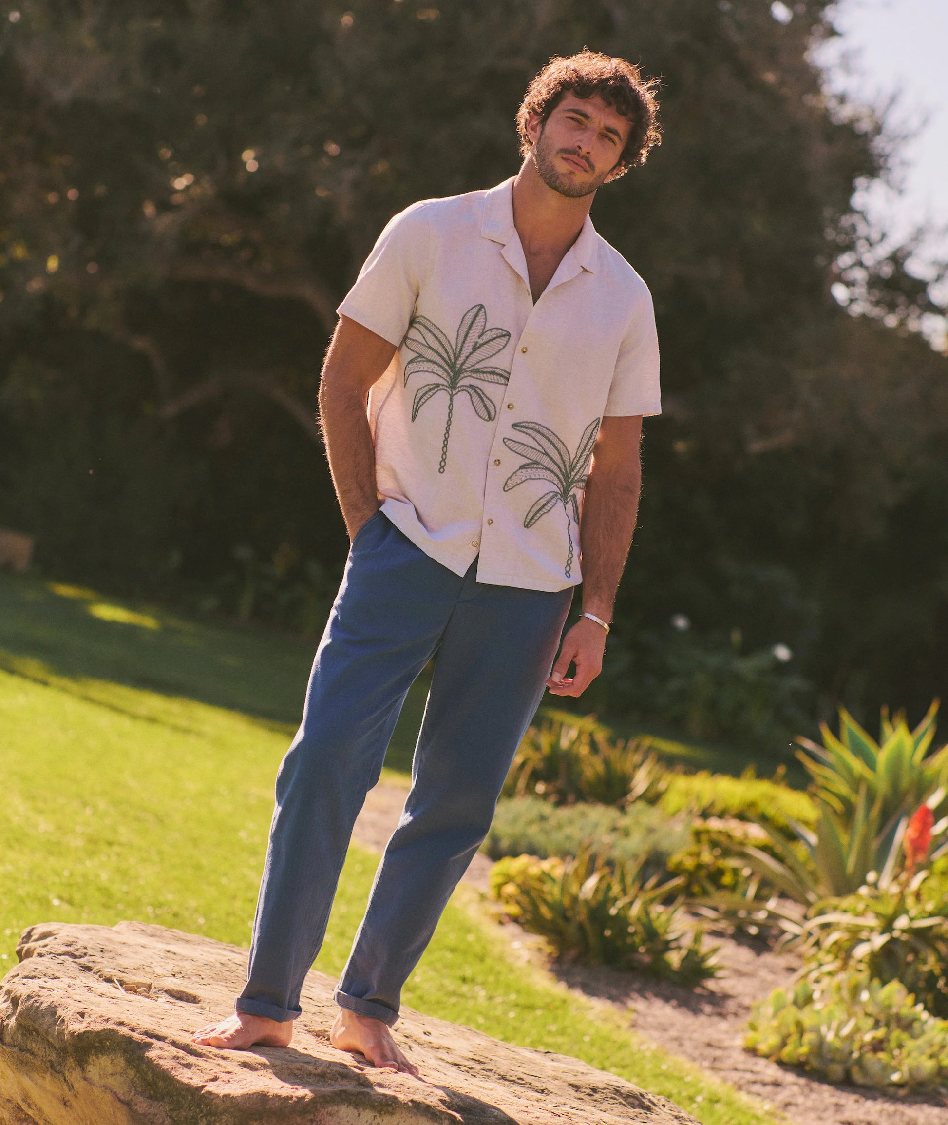 Man standing on a rock in a garden wearing a white shirt with palm tree designs and blue pants.