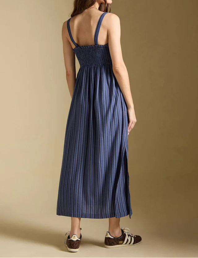 A woman shown from the back wearing a blue pleated midi dress with thin shoulder straps and a smocked bodice, paired with a matching blue cap and brown sneakers, against a solid beige studio background.