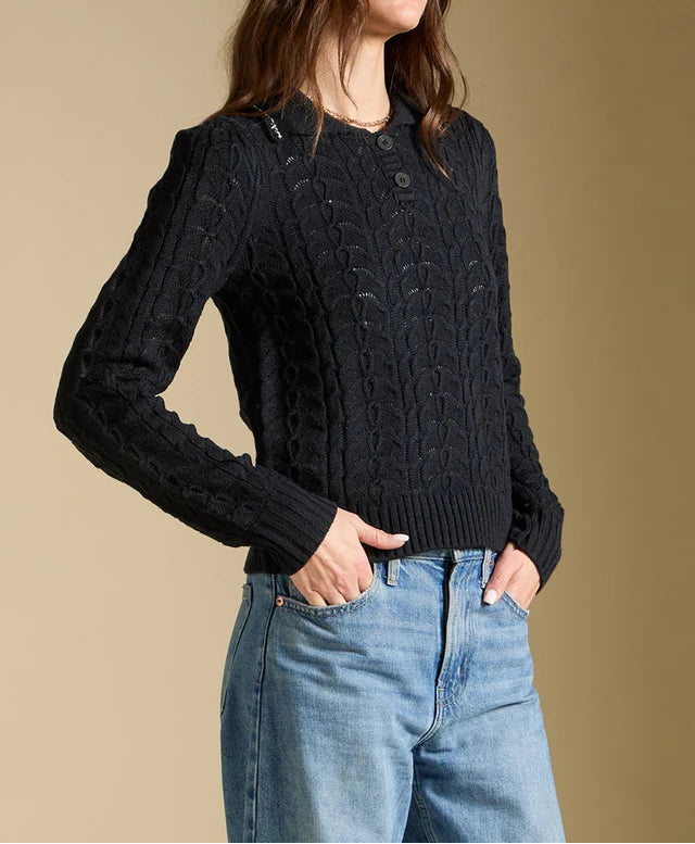Woman wearing a black cable knit sweater and blue jeans against a beige background